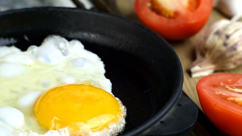 Pouring chicken egg into hot skillet and cooking fried eggs. Tomatoes and garlic. Scrambled eggs cooking. Simple tasty food preparation concept.