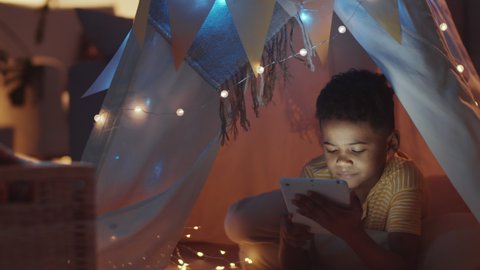 Tilt down shot of happy little African American boy lying in teepee decorated with lights, playing on digital tablet and then looking at camera with smile in dark kids room