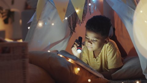 Smiling African American boy lying in handmade teepee tent decorated with lights, holding flashlight and reading book in the evening at home