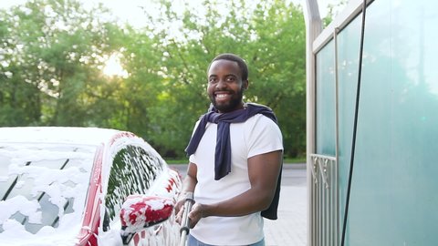 Good-looking joyful 30-aged black-skinned guy with beard in casual clothes posing nearby his lux class auto with high pressure washer in self-service car wash