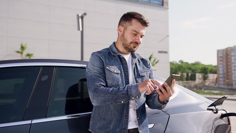 Attractive smiling stylish bearded guy leans on his modern black electrocar that plugged in specially-equipped charging station and watching funny videos on mobile
