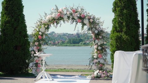 Place for bridal ceremony in white color against the background of the river. Wedding arch with bouquets of pastel roses. Slow motion.