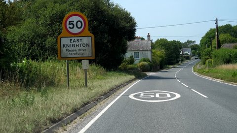 Car point of view driving past East Knighton village name board and speed restriction sign in Dorset, England on a sunny summer day.