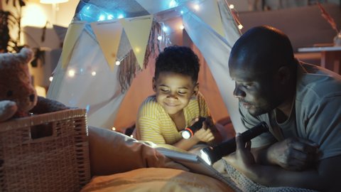 Joyous African American dad and little son holding flashlights, reading book, chatting and laughing while he lying in teepee tent decorated with lights in dark room in the evening
