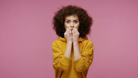Insecure frightened young woman afro hairstyle in hoodie biting nails, feeling worried nervous about serious troubles, stress and anxiety disorder concept. indoor studio   isolated on pink background