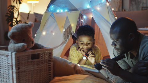 Happy African American father holding flashlight and reading fairy tale to cheerful little son while he lying in teepee tent decorated with lights in dark room at night