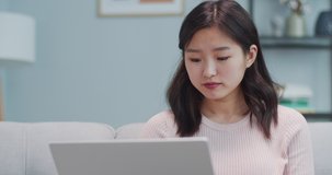 Charming Asian woman is sitting on sofa and studying in living room. Smart young female student using laptop computer to read information. Education, distance learning, Interner source.