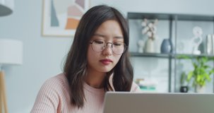 Good-looking Asian woman in glasses sitting at table and working in office room at home. Hardworking female worker shutting laptop computer, looking at camera and smiling.Business, enterprenuership.