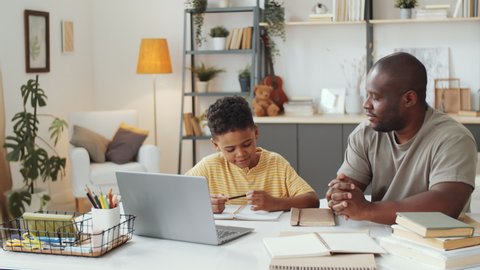 African American father sitting with little son at table in the living room, pointing on laptop screen and explaining homework to kid as he taking notes