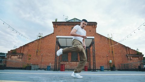 Young attractive caucasian man dancing hip hop on the roof with modern stage on background. Professional male dancer training alone enjoying freestyle dance. Artistic performance show online. 4K