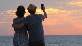 Couple making selfie. Romantic sunrise sunset background. Silhouettes of travelers on a journey looking at the sunrise on the beach. Two lovers making selfie video photo on beach vacation. Back view