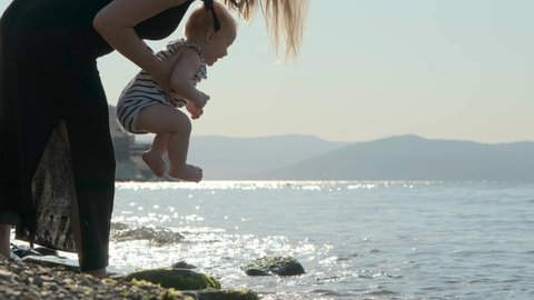 Shooting against the sun. Close up slender mother in a black dress with her daughter in her arms. The mother puts the baby's feet into the sea or ocean. Happy family is having fun.