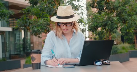 Serious female entrepreneur writing down to do list to achieve her goals. Woman working in the outdoor business park on laptop. Business woman is planning agenda of professional business meeting, 4K. 