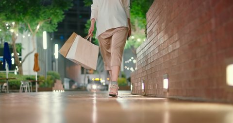 4K slow motion shopping video. Front view of woman in evening silk dress on fashionable heels walking at city park carrying shopping bags on beautiful summer night with street lights on background.