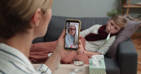 Mother holding smartphone and talking with elderly female doctor. Woman having online medical consultation while her child son lying on sofa with thermometer. Concept telemedicine.