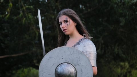 A portrait of a medieval woman warrior with combat make-up, dressed in chain mail armor with a shield and a sword in her hands, stands in a combat stance against a background of forest bokeh. The wind