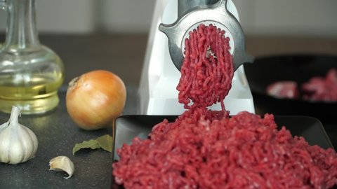 Preparation of minced fresh beef through electric meat grinder on background with onion, garlic and olive oil. Mans hands put meat in meat-mincer. Food background
