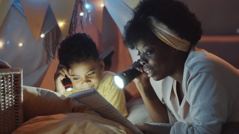 Cheerful African American mother and little son lying in teepee tent decorated with lights, holding flashlights and reading fairytale story aloud while spending evening together at home.