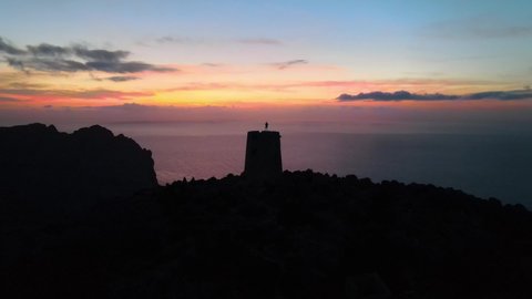 Aerial shot flying over the silhouette of a man standing on top of a watchtower on a spectacular sunset