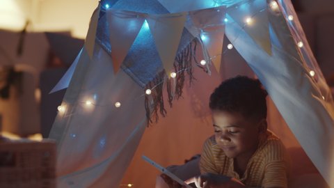 Cheerful little African American boy lying in cozy teepee decorated with lights and browsing the Internet on digital tablet in dark room at night.