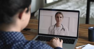 Therapist cardiologist provide medical heart disease consultation to client by video call, laptop screen view over patient shoulder sit at desk got help from woman practitioner doctor in white coat