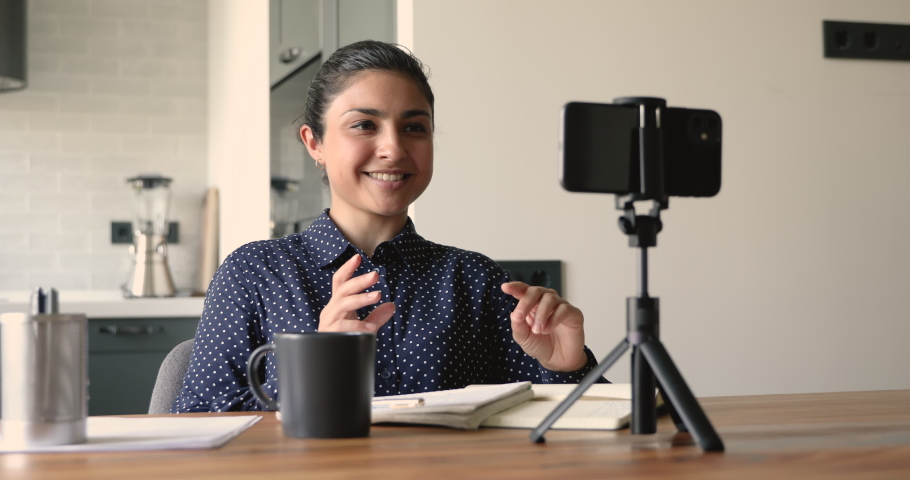 Indian woman sit at desk put phone on tripod record video vlog, vacancy applicant films job interview tell about education qualification feels confident. Modern tech, influencer work activity concept Royalty-Free Stock Footage #1058470687