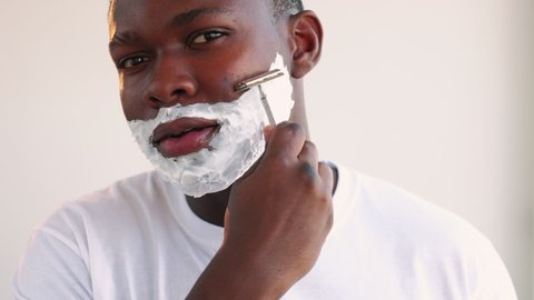 Man grooming. Face hygiene. Portrait of confident black guy in white t-shirt shaving beard with foam razor isolated on light copy space background. Morning routine.