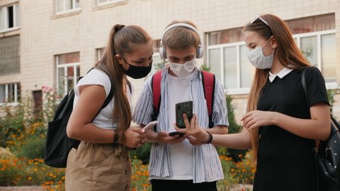 Schoolchildren wearing protective medical masks communicate in the school yard, use smartphones. Education during the coronavirus pandemic