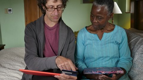 Caucasian man teaching an elderly black woman how to buy something on her tablet with a credit card