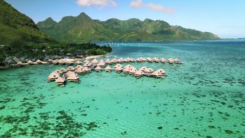 Drone aerial shot 4k. Overwater bungalows resort in coral reef lagoon ocean by beach. Paradise getaway in Moorea, French Polynesia, Tahiti. View from above glide shot.