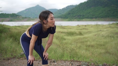Tired young Asian woman take deep breathe after running in a meadow. Sweaty athletic female training cardio jogging on grass field trail. Outdoor workout lifestyle and extreme sport training concept.