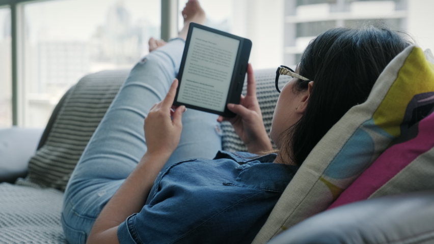 Happy female student reading ebook and lying on couch. Young woman relaxing with book. Caucasian girl and lifestyle, leisure and relaxation at home Royalty-Free Stock Footage #1058486716