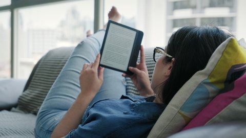 Happy female student reading ebook and lying on couch. Young woman relaxing with book. Caucasian girl and lifestyle, leisure and relaxation at home
