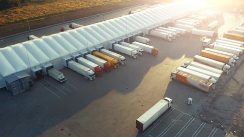 Aerial view of the logistics park with warehouse and loading hub. A lot of semi trucks with cargo trailers awaiting for loading/unloading goods at sunset
