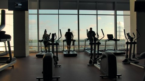 Silhouettes of three unrecognizable people running on treadmills in an empty gym against a large window on a sunny day. eft to right extreme slow motion shot.
