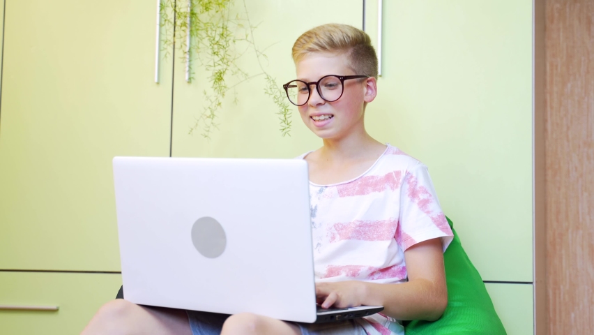 Cute little smiling boy use laptop and wave to someone on video call. Teen with glases uses laptop in home. Waving hand at the webcam. 