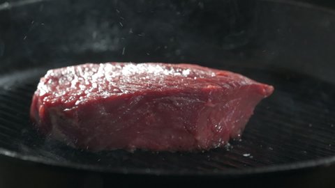 Chunk of raw steak is dropped into a hot grill pan. Close-up of falling raw beef steak on the grill pan and smoke on the background.