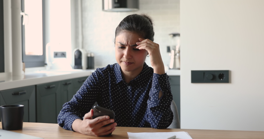 Indian ethnicity woman sit at desk holding smart phone feels unhappy negative emotions upset by receiving terrible news by sms. Device damaged, lost internet connection, need gadget fix repair concept | Shutterstock HD Video #1058489860