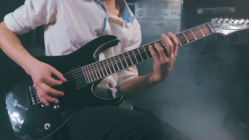 Medium Close-Up Shot of a Handsome Young Man Playing Electric Guitar in a Dark Smoky Room. Active Emotional Musician in a White Shirt is Recordind Music at the Home Studio Against the Black Background