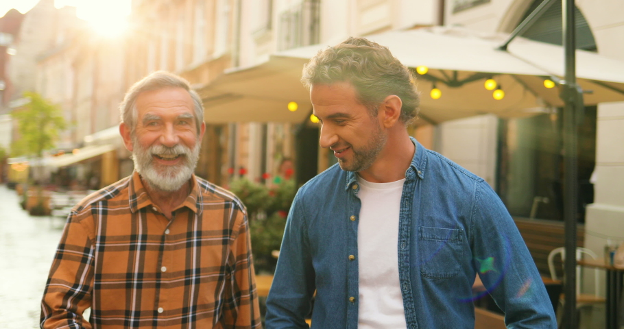Two male generations walking the street in town and talking. Father and son strolling in city and talk with smiles outdoor. Senior dad sharing his wisdom with young man outdoor. Men of different ages.