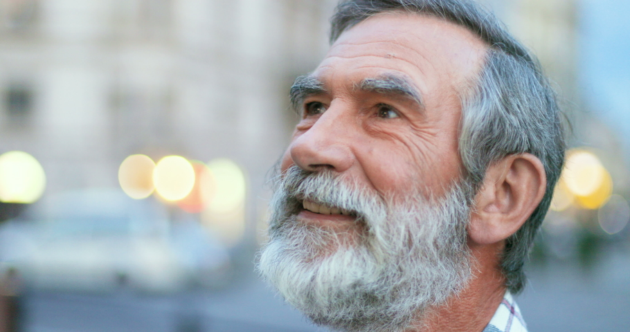 Close up of face of senior Cauasian man with gray hair and beard. Portrait of handsome old grandfather looking at side ad then turning face to camera with smile at street in town. Outdoors. | Shutterstock HD Video #1058490907