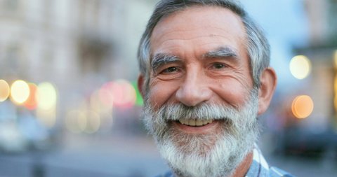 Close up of face of senior Cauasian man with gray hair and beard. Portrait of handsome old grandfather looking at side ad then turning face to camera with smile at street in town. Outdoors.