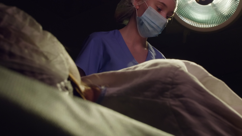 Low angle shot of female nurse in medical uniform, hat, mask and gloves giving newborn baby to mother in dark delivery room with surgical light Royalty-Free Stock Footage #1058491687
