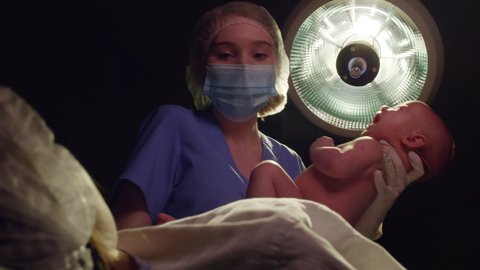 Low angle shot of female nurse in medical uniform, hat, mask and gloves giving newborn baby to mother in dark delivery room with surgical light