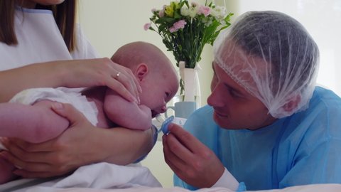 Close up shot of father in medical gown and disposable hat giving soother to crying newborn baby while mother holding child on arms and caressing in hospital ward
