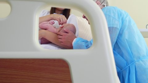 Tilt up shot of happy man in medical gown and hat kissing and caressing newborn baby on arms of his wife while visiting her after childbirth in hospital ward