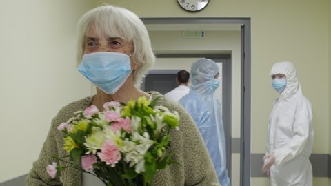 Senior woman in medical face mask waving to doctor and nurses in protective uniforms, then walking with flowers through corridor while leaving hospital after covid-19 recovery