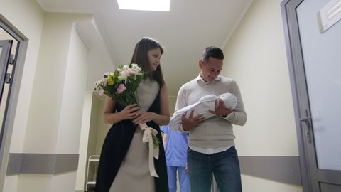 Delightful husband meeting his wife in maternity hospital and taking newborn baby while cheerful nurse giving bouquet of flowers to woman. Loving parents walking together through hallway