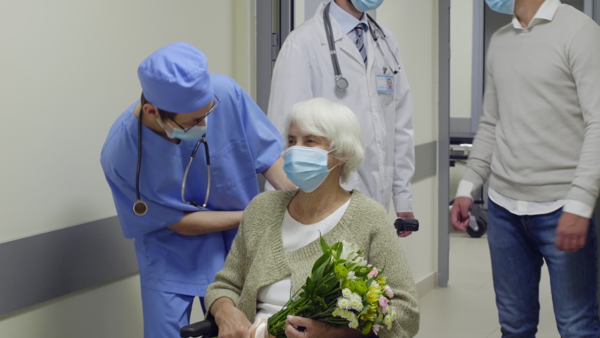 Tilt up of elderly woman in medical face mask holding flower bouquet while leaving hospital after recovery; her son giving handshake to doctor and pushing wheelchair | Shutterstock HD Video #1058491834