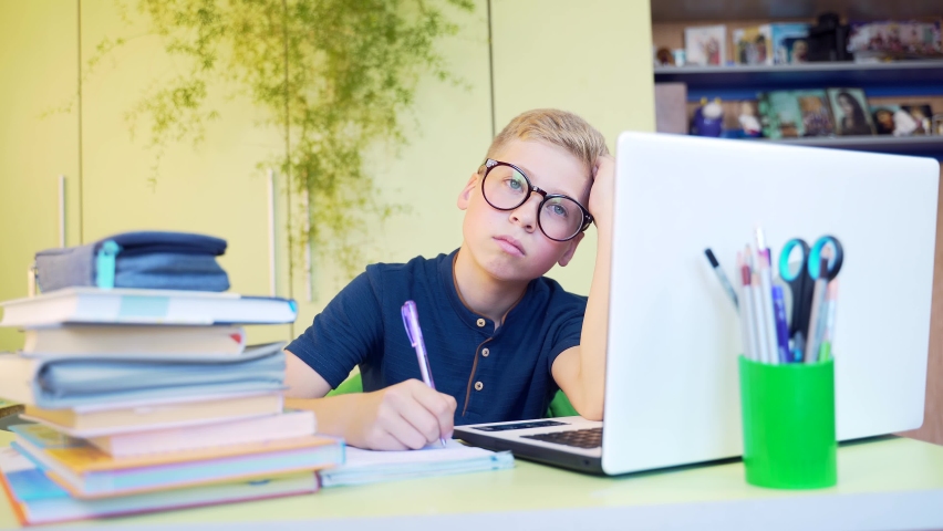 Cute tiring teenage boy sitting at the table and writing at notebook during distance learning at home, with eyeglasses. Boring child, schoolboy during online studing with books and white laptop.